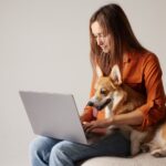 Woman working on laptop with Corgi dog on lap, cozy remote work lifestyle.